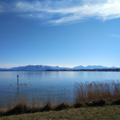 Klarer Himmel über einem ruhigen See mit Bergen im Hintergrund und Schilf am Ufer.